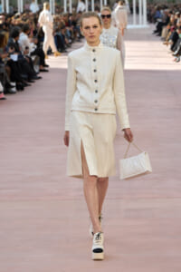 Model walking a runway in a white textured suit with a matching skirt and jacket, carrying a small white quilted handbag with a gold chain.