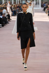 Model struts down a runway in a black textured suit with a white cape draped over the shoulders and cream platform shoes.