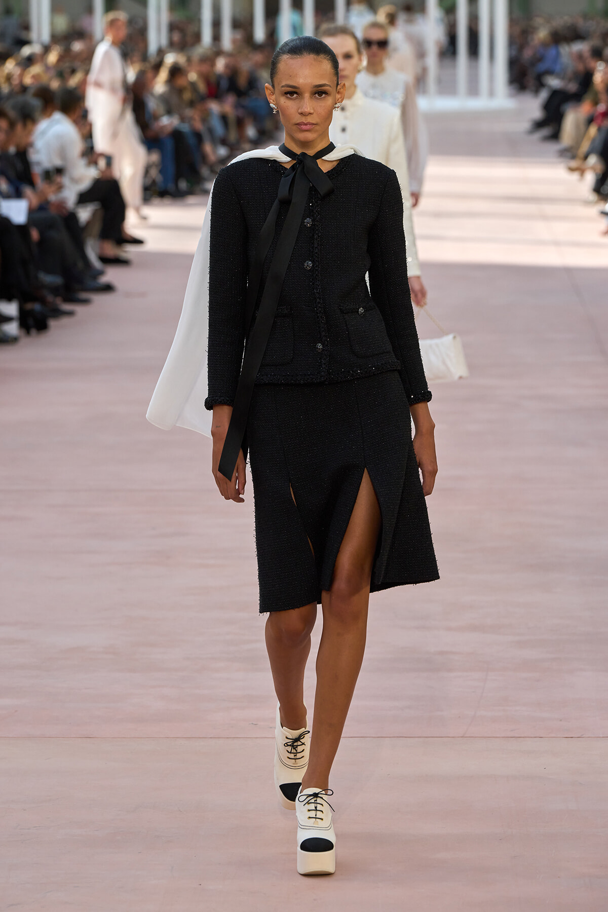 Model struts down a runway in a black textured suit with a white cape draped over the shoulders and cream platform shoes.