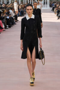 Model on a runway wearing a black textured dress with a white collar, gold platform shoes, and a black handbag with a gold chain.