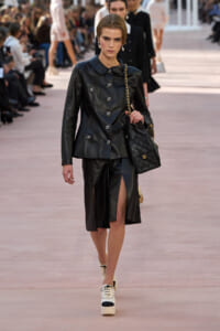 Model walks the runway in a black leather jacket and skirt with a front slit, carrying a quilted black handbag with a gold chain.