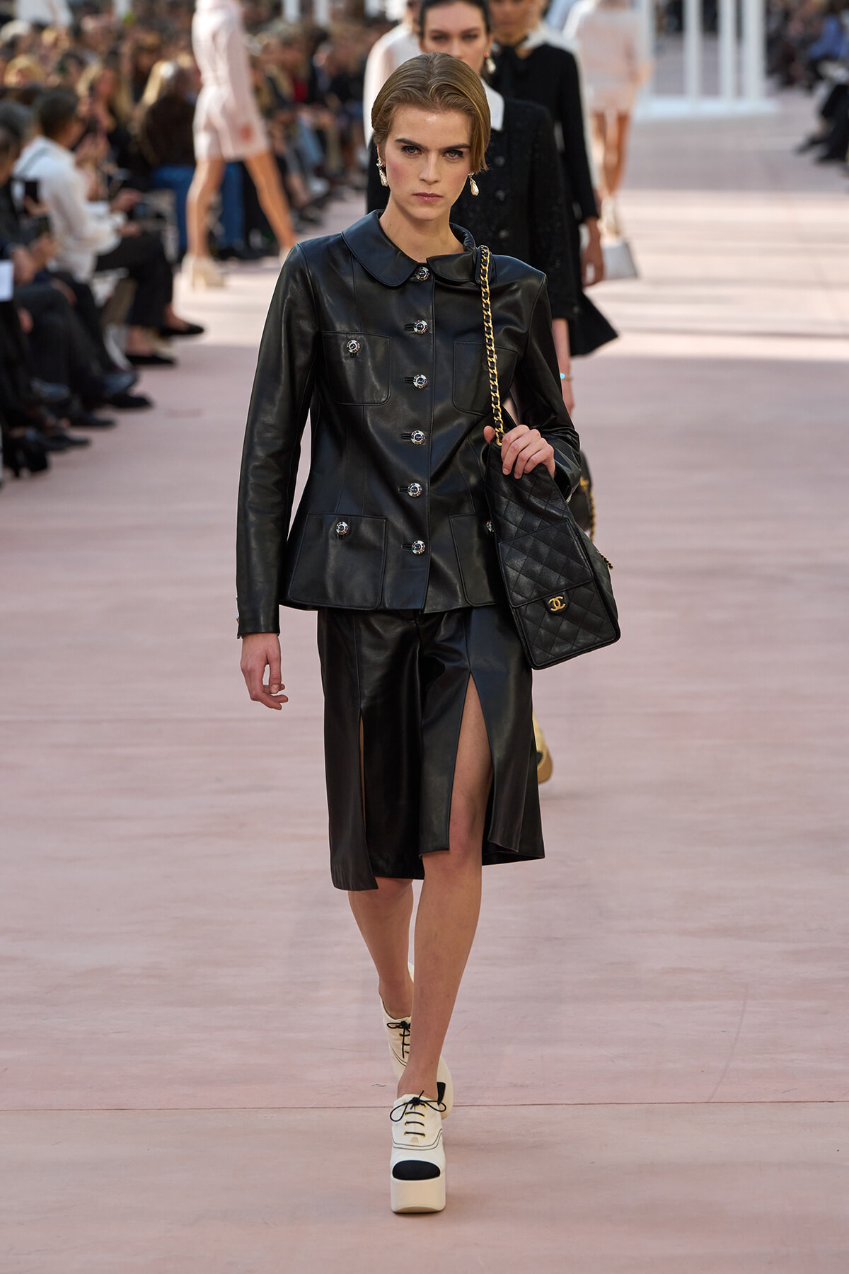 Model walks the runway in a black leather jacket and skirt with a front slit, carrying a quilted black handbag with a gold chain.