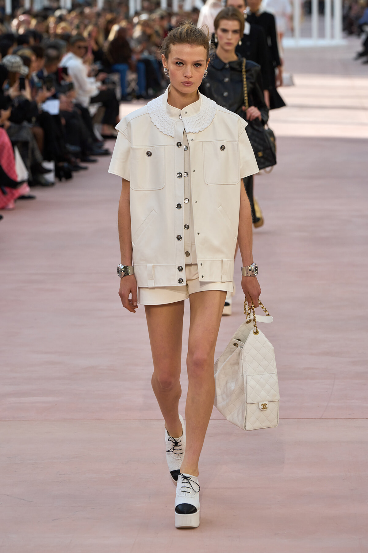Female model walking a runway in an all-white outfit: short shorts, button-front jacket with oversized pockets, quilted white handbag with gold chain.