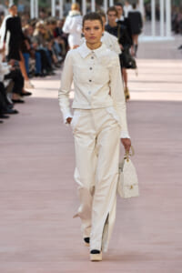 Model walking a runway in an ivory-white suit with a large fabric flower at the shoulder, holding a quilted white handbag with a gold chain.