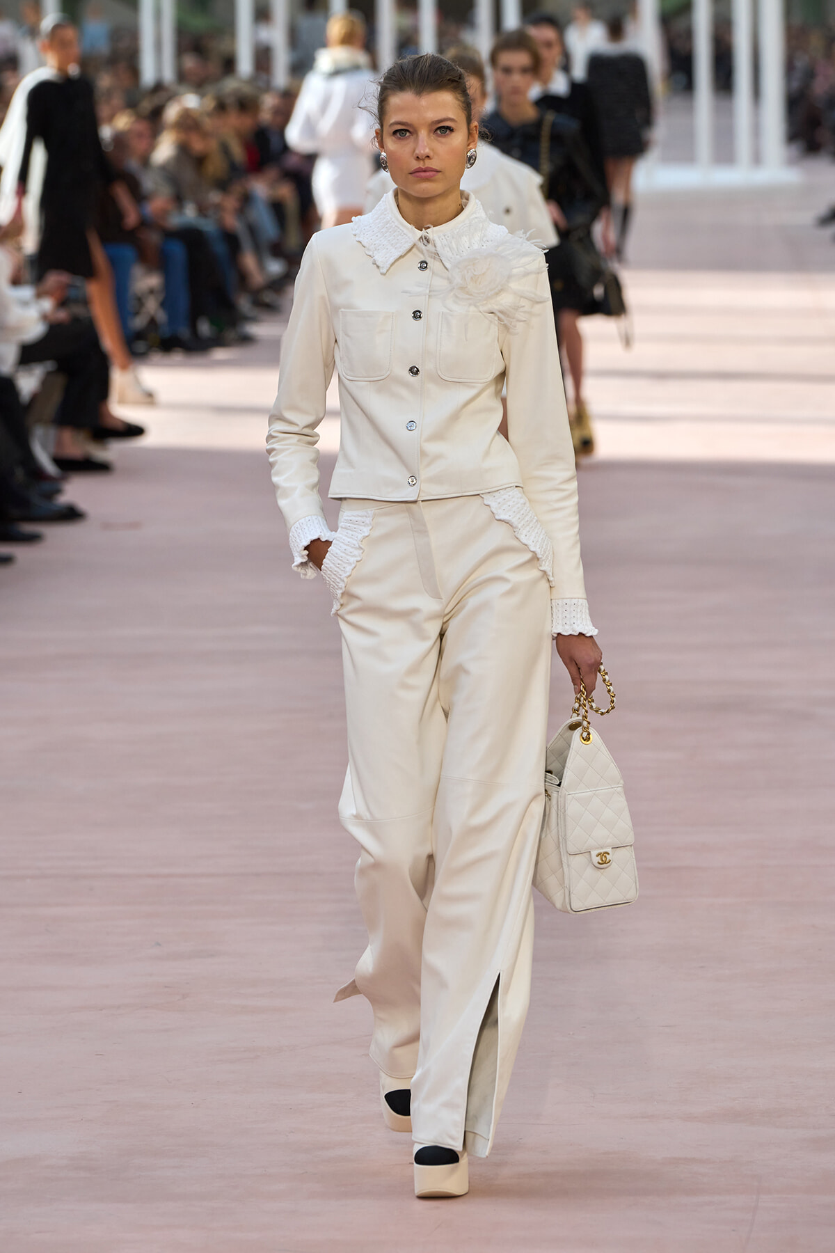 Model walking a runway in an ivory-white suit with a large fabric flower at the shoulder, holding a quilted white handbag with a gold chain.