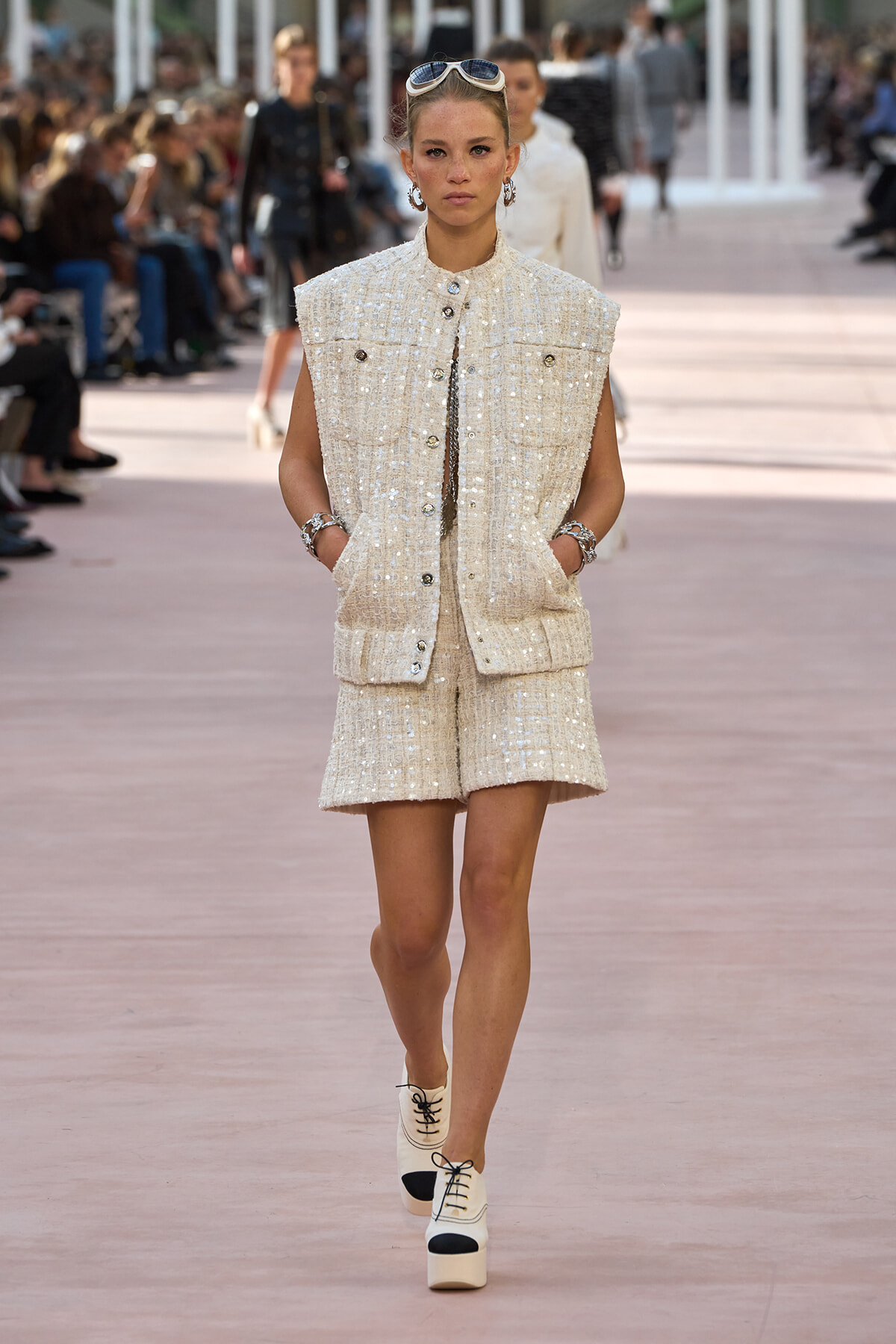 Runway model in a cream tweed sleeveless vest and matching shorts with sunglasses on her head, walking the catwalk in platform shoes.