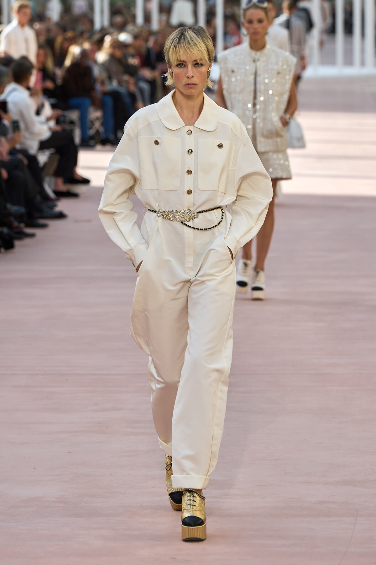Model walks a fashion show wearing a cream jumpsuit with a belted waist and gold platform boots on a pink runway, audience in the background.