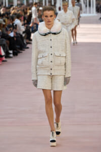 Model walks the runway in a cream tweed jacket and matching shorts with a fluffy pastel collar and gloves.