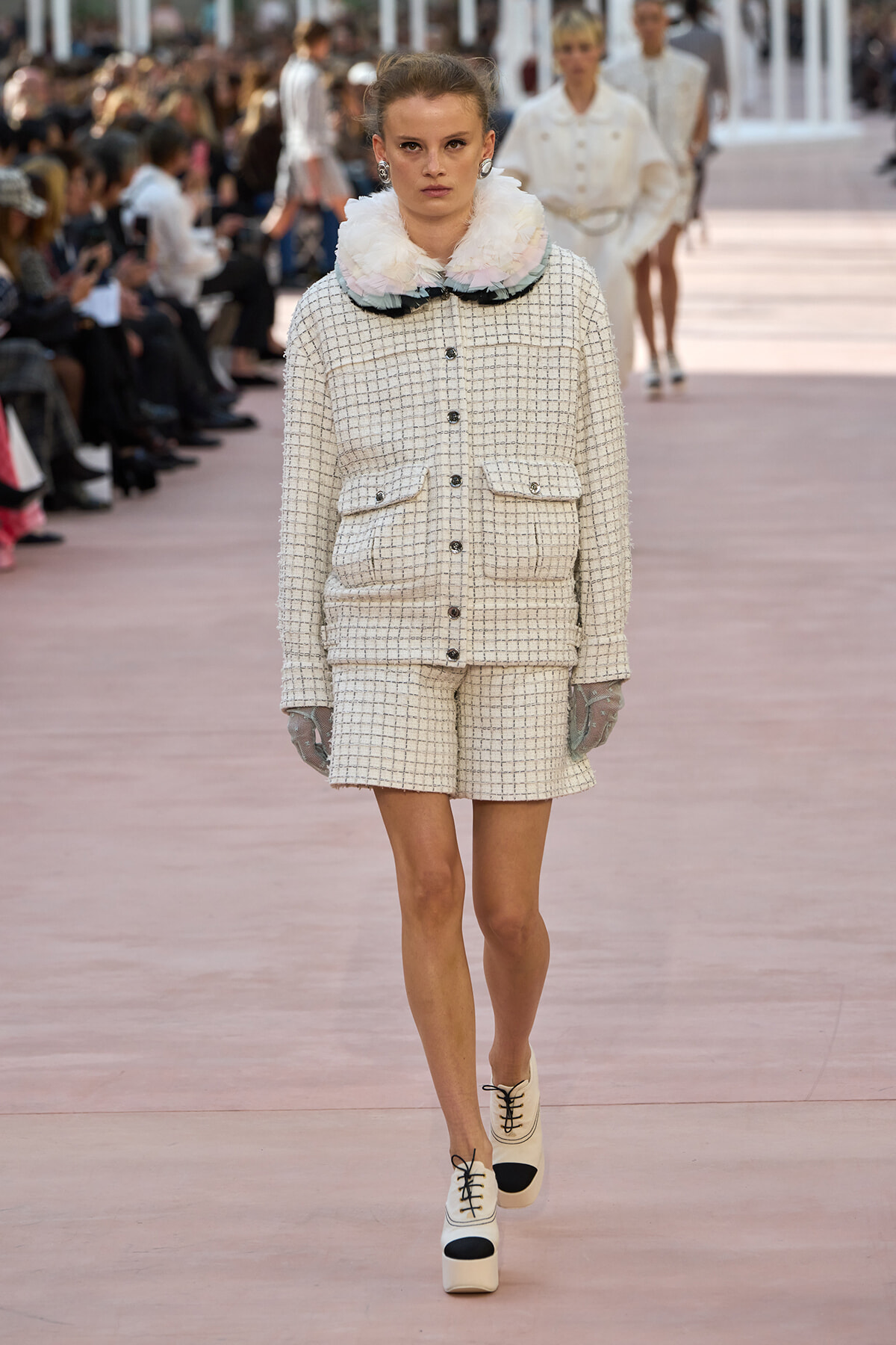 Model walks the runway in a cream tweed jacket and matching shorts with a fluffy pastel collar and gloves.
