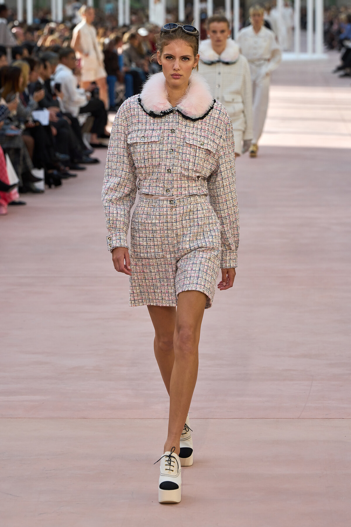 Model walking a fashion show runway in a pastel tweed matching set with pink fur collar and white platform shoes, sunglasses on head.