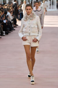 Female model walks the runway in a pastel tweed jacket and matching shorts, with a fluffy scarf at the neck.