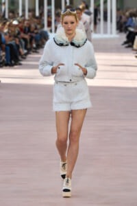 Female model walking a runway in a pale blue textured jacket and matching shorts with a large ruffled collar, hands in pockets, sunglasses on head.