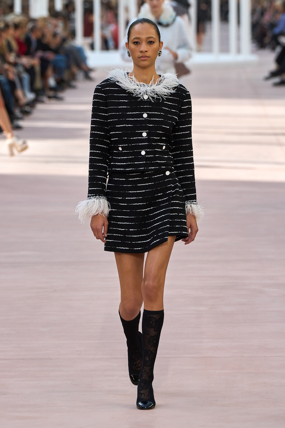 Model walks runway in a black-and-white striped dress with white feather trim at the collar and cuffs, paired with knee-high lace socks.