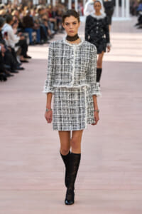 Runway model in a black-and-white tweed two-piece outfit with frayed white trim, pairing a jacket and skirt, plus black lace stockings and boots.