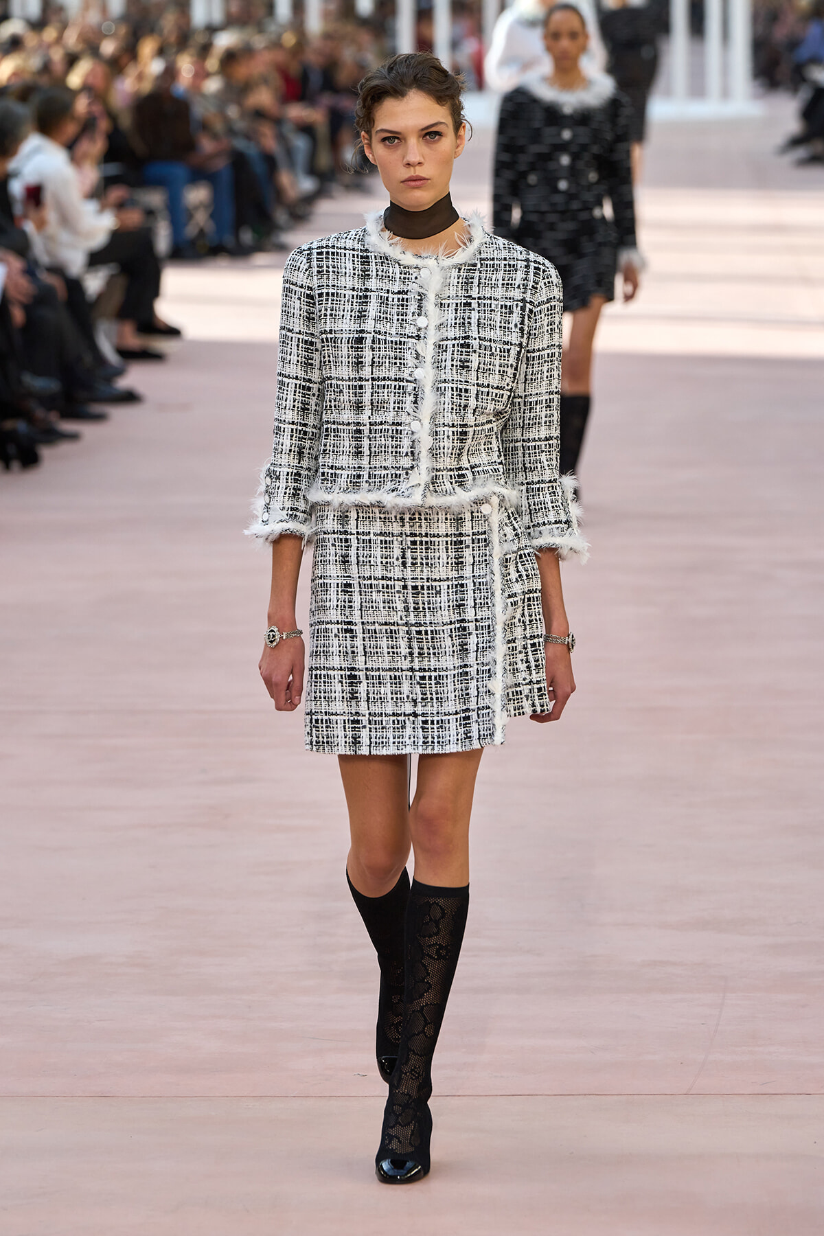 Runway model in a black-and-white tweed two-piece outfit with frayed white trim, pairing a jacket and skirt, plus black lace stockings and boots.