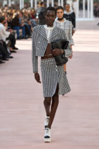 Model in a black-and-white checkered cropped blazer and matching skirt, white blouse, carrying a black clutch on runway.