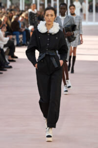Female model walks a runway in a black belted jumpsuit with a white feather collar; audience out of focus in background.