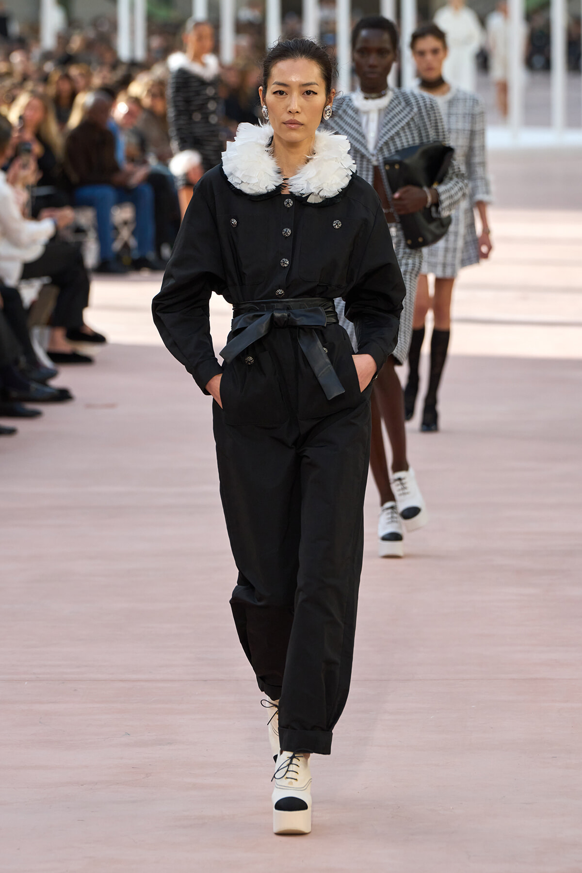 Female model walks a runway in a black belted jumpsuit with a white feather collar; audience out of focus in background.