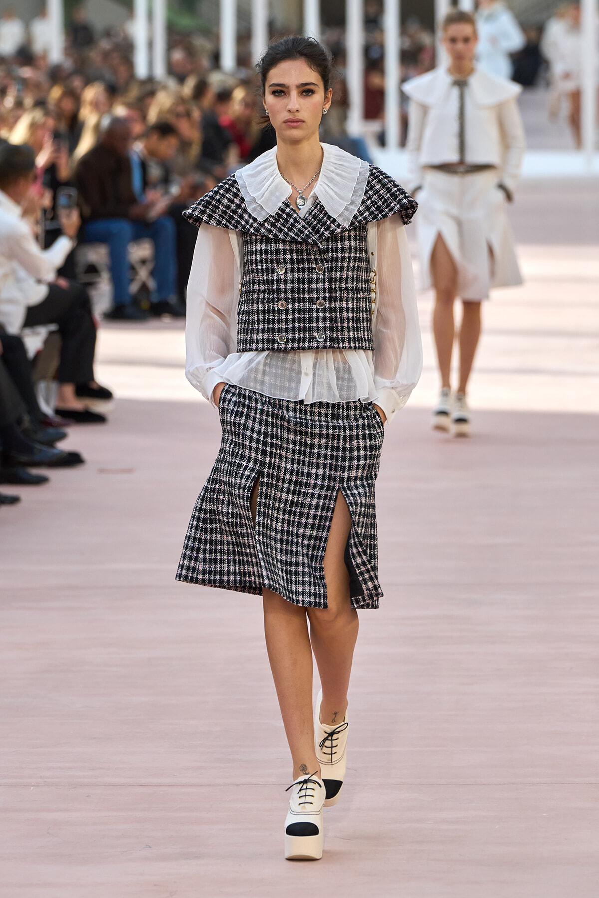 Model walking a fashion runway in a black-and-white plaid two-piece suit with a sheer blouse and oversized white collar; skirt has front slits and she wears two-tone platform shoes. Audience blurred in background.