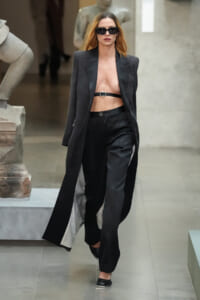 Female model walking a runway in a black oversized blazer, high-waisted black pants, and a thin belt-bra top, with dark sunglasses.