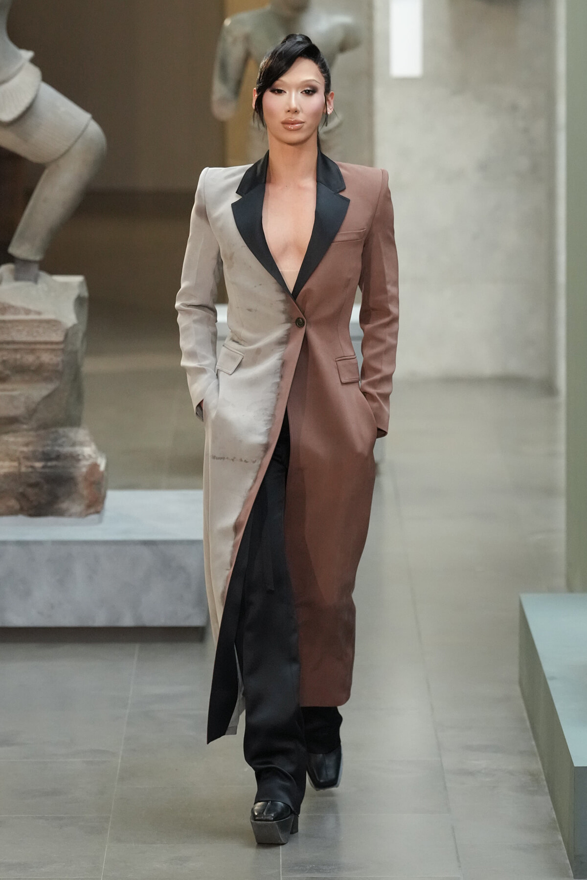 Female model walking the runway in a color-blocked long coat split beige and brown with black lapels, deep V neckline, and black pants with platform boots.