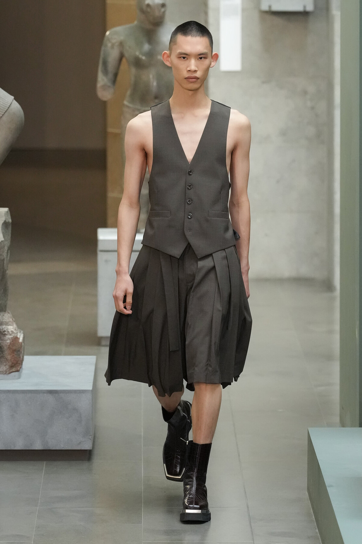 Male model walking a runway in a charcoal gray vest with a V-neck over a matching pleated skirted bottom and black boots.