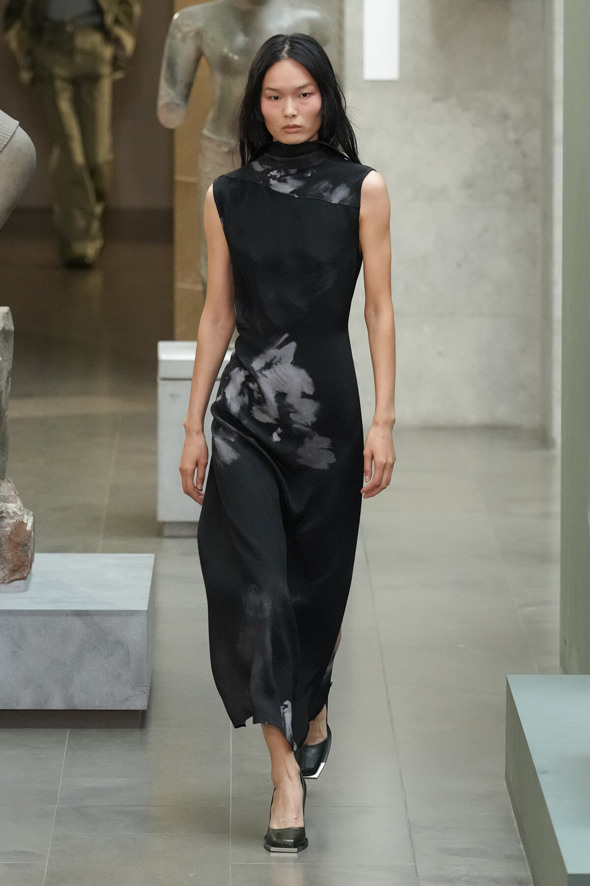 Model walking a runway in a sleeveless black dress with a gray abstract print and black shoes in a modern gallery setting.