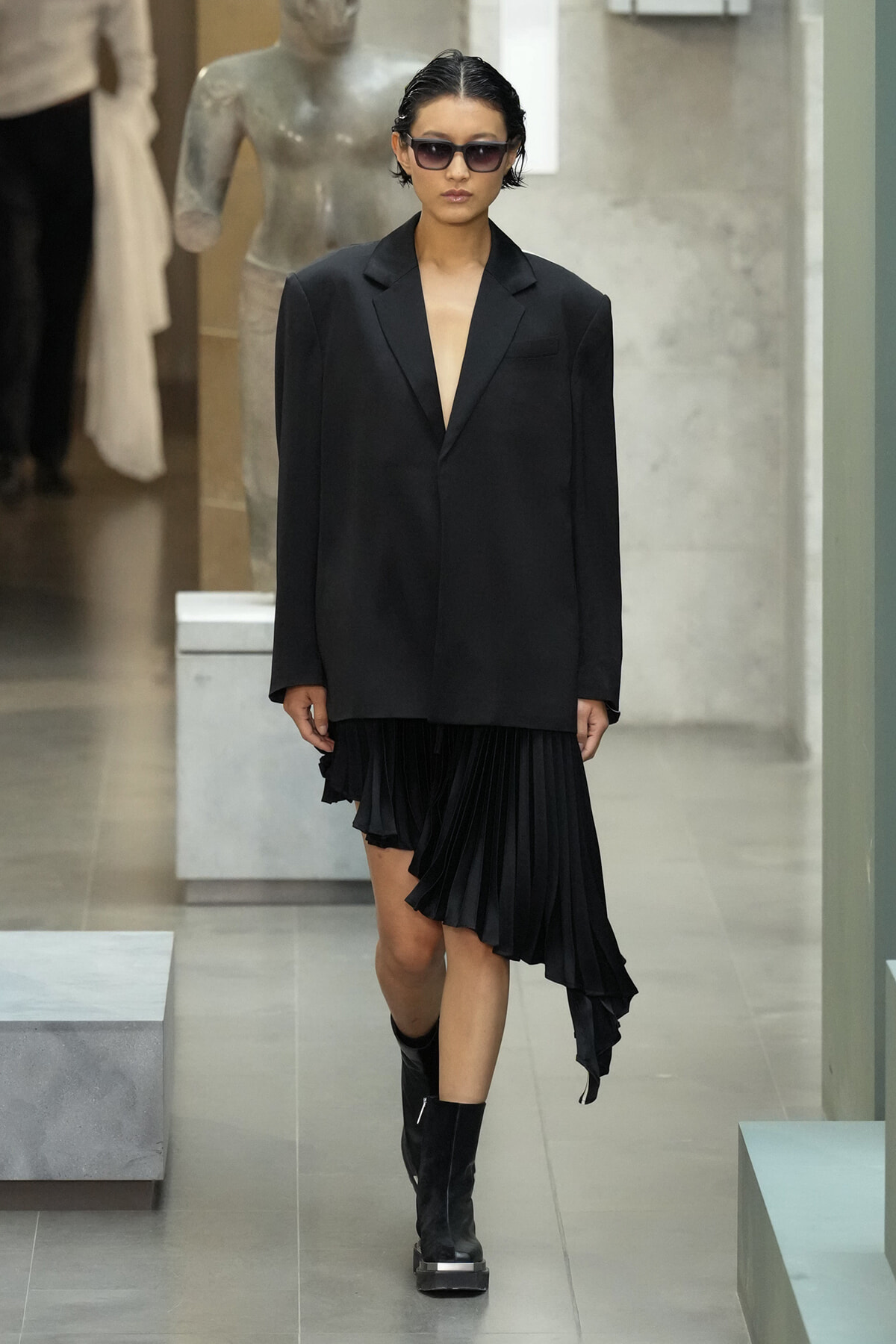Model in an oversized black blazer with a black asymmetric pleated skirt, wearing sunglasses and black boots, walking down a gallery-like runway.
