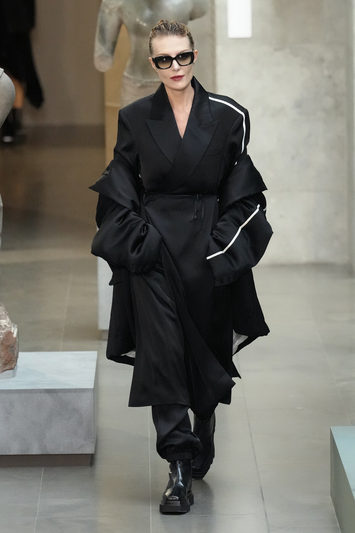 Model walking a runway in a long black belted coat with exaggerated sleeves and white piping, wearing sunglasses and boots.