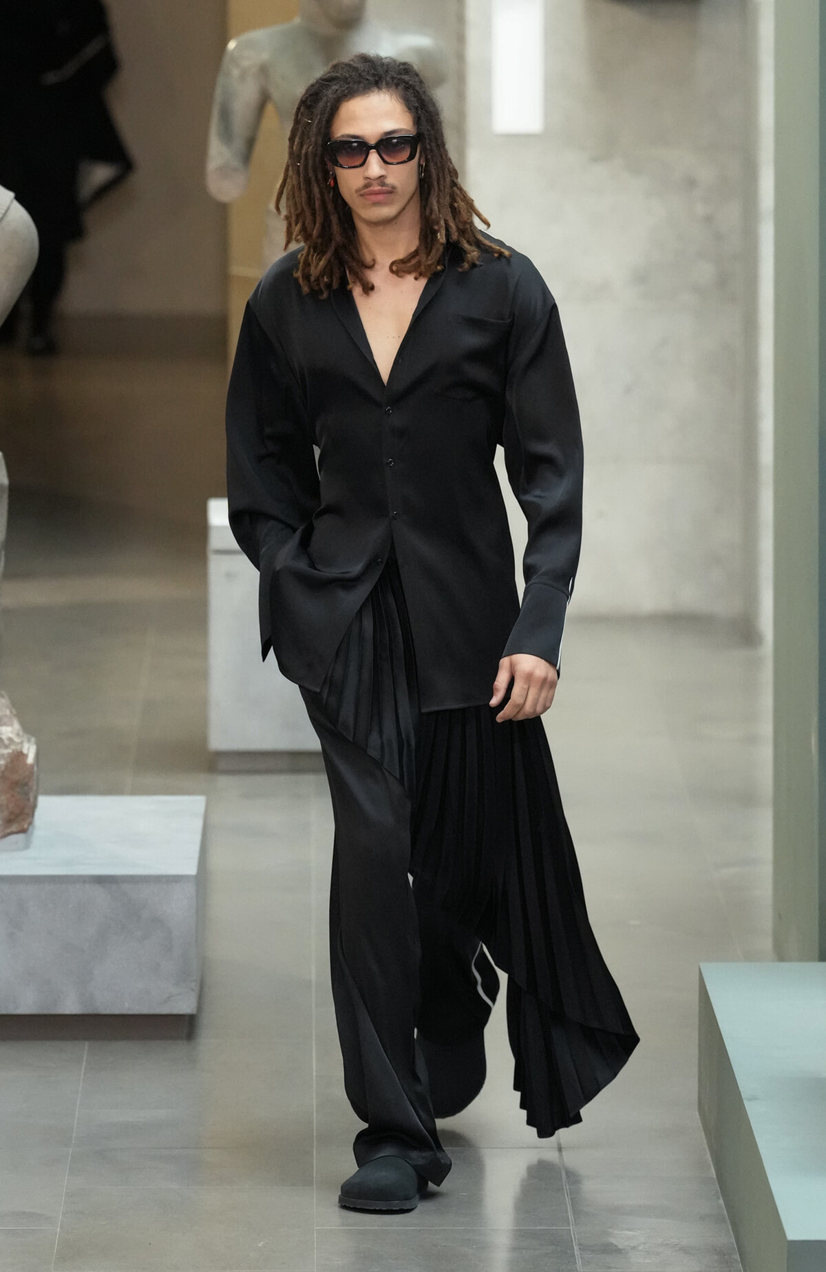 Male model with dreadlocks in all-black avant-garde outfit on a runway, wearing sunglasses.