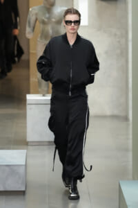 Model wearing a black bomber jacket and black cargo pants with white piping, sunglasses, walking in a gallery-like space.