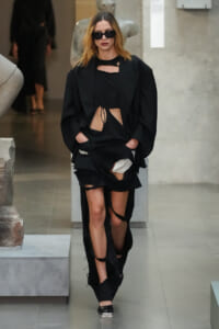 Model walking in a black, distressed ensemble: oversized blazer over a torn black dress with multiple rips, wearing dark sunglasses on a runway.