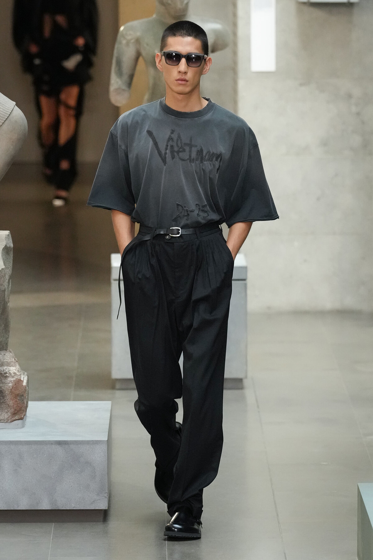 Male model walking a runway in a loose dark gray graphic T-shirt, black high-waisted pleated pants, belt, and black sunglasses.