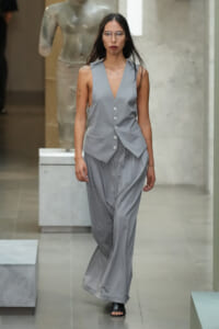 Female model walks a runway in a light gray sleeveless vest with a deep V, matching wide-leg pants, and clear protective glasses.