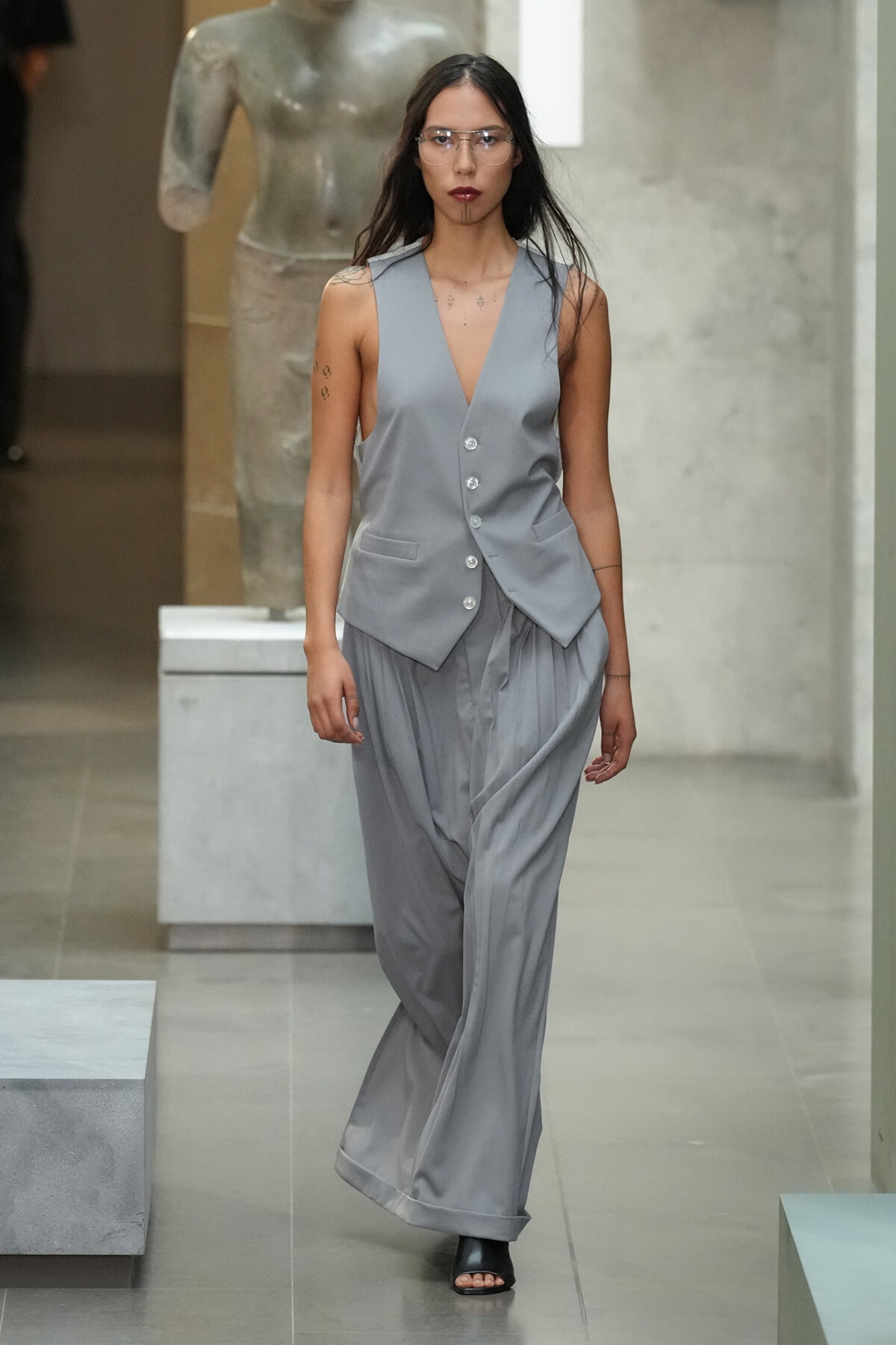 Female model walks a runway in a light gray sleeveless vest with a deep V, matching wide-leg pants, and clear protective glasses.