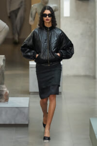 Model in a glossy black oversized puffer jacket, black layered skirt, and sunglasses walking indoors in a fashion setting.