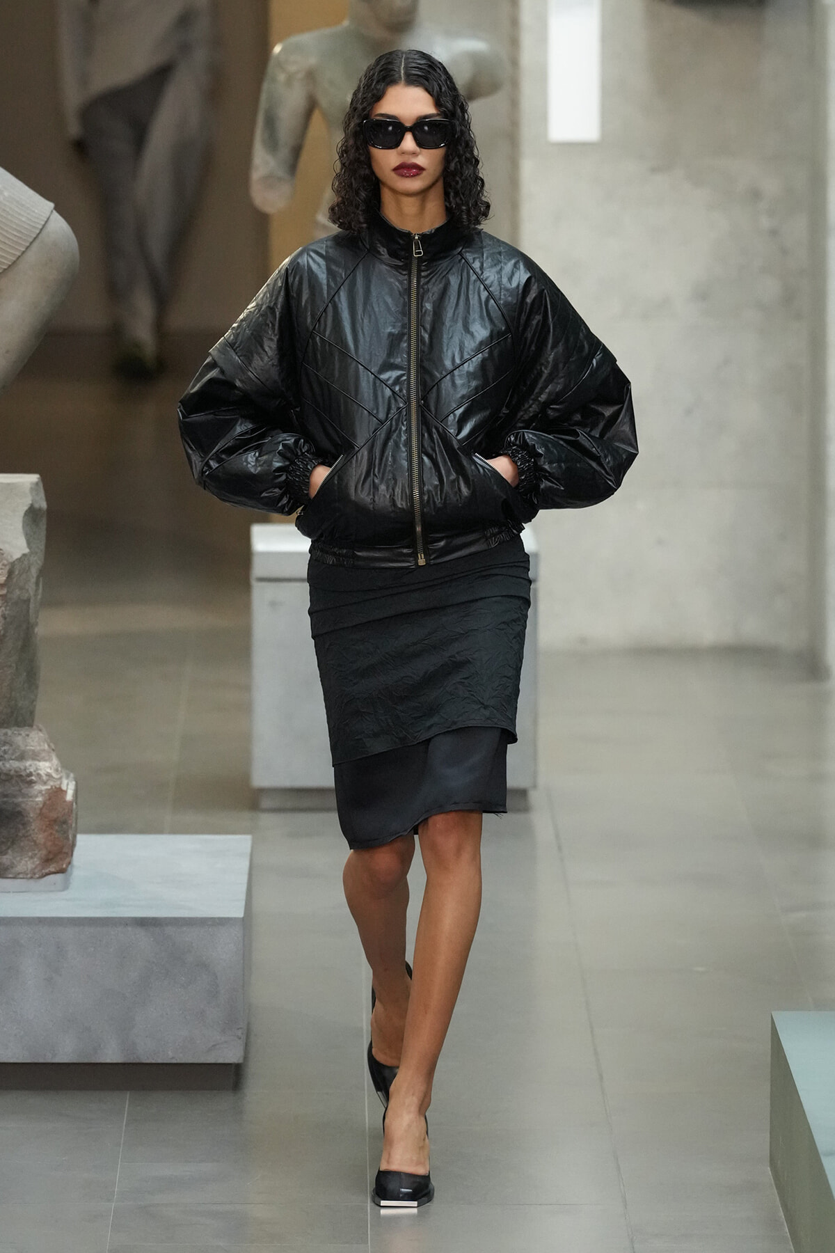 Model in a glossy black oversized puffer jacket, black layered skirt, and sunglasses walking indoors in a fashion setting.