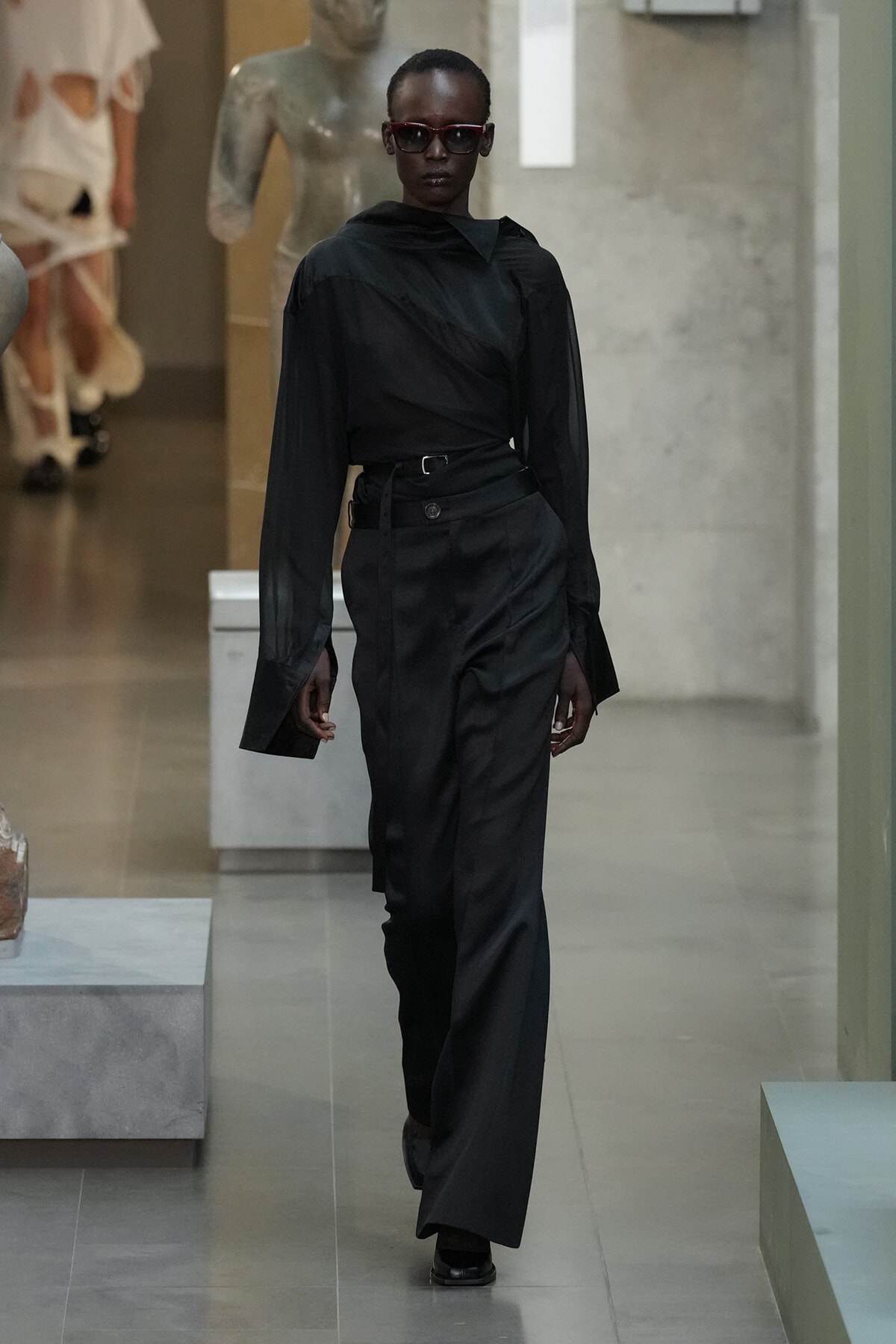 Model wearing an all-black satin outfit with wrapped top and wide-leg pants, red sunglasses, walking down a gallery runway.