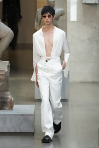 Model in a white, plunging blouse and high-waisted pants with a belt, wearing sunglasses and black shoes, walking in a gallery.