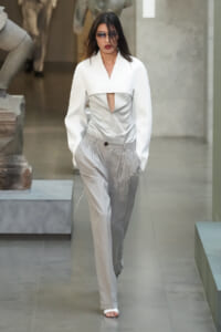 Model walking the runway in a white cropped top with a triangular cutout over a silver-gray bodice, paired with high-waisted pleated gray trousers and sunglasses.