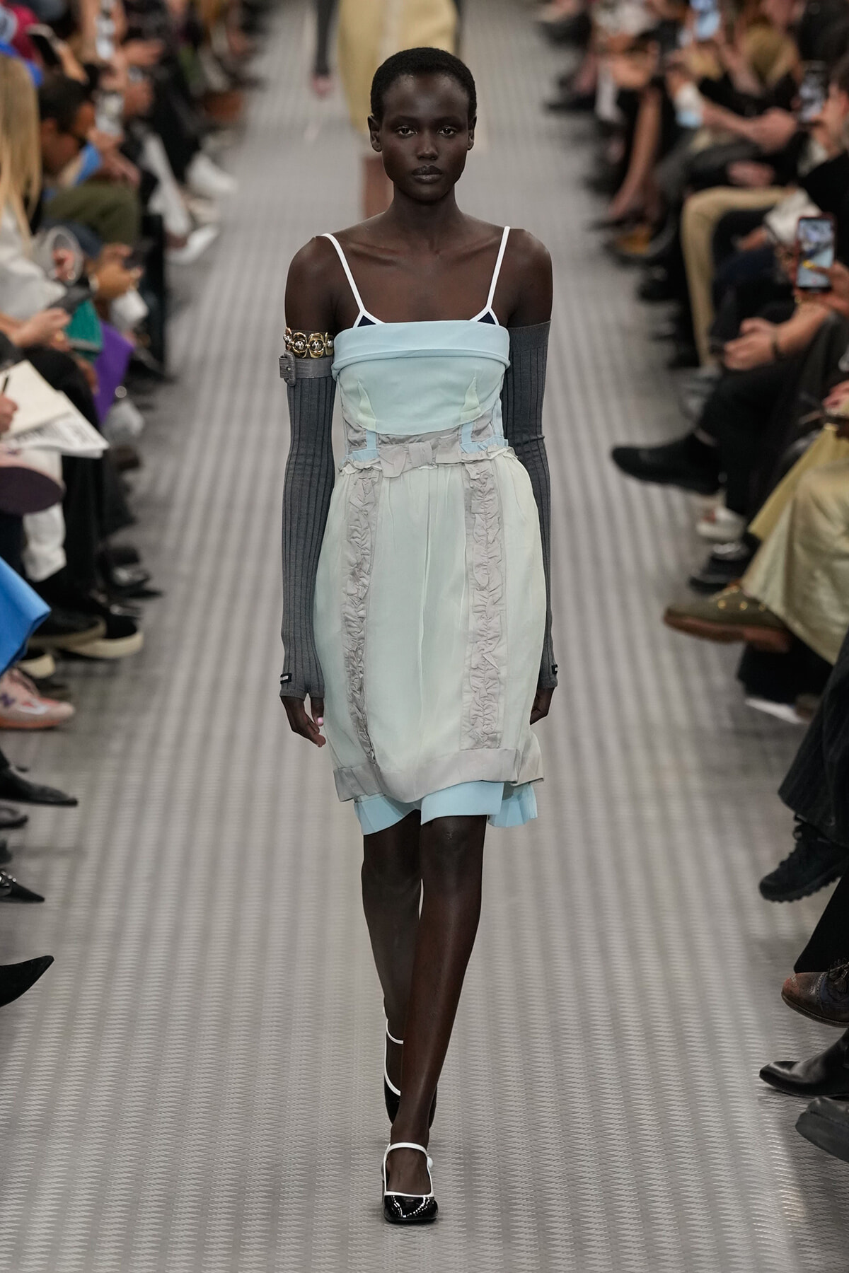 Model walks a runway in a pale blue strapless dress with ruched panels and a ruffled skirt, paired with gray elbow-to-wrist sleeves and black-and-white heels