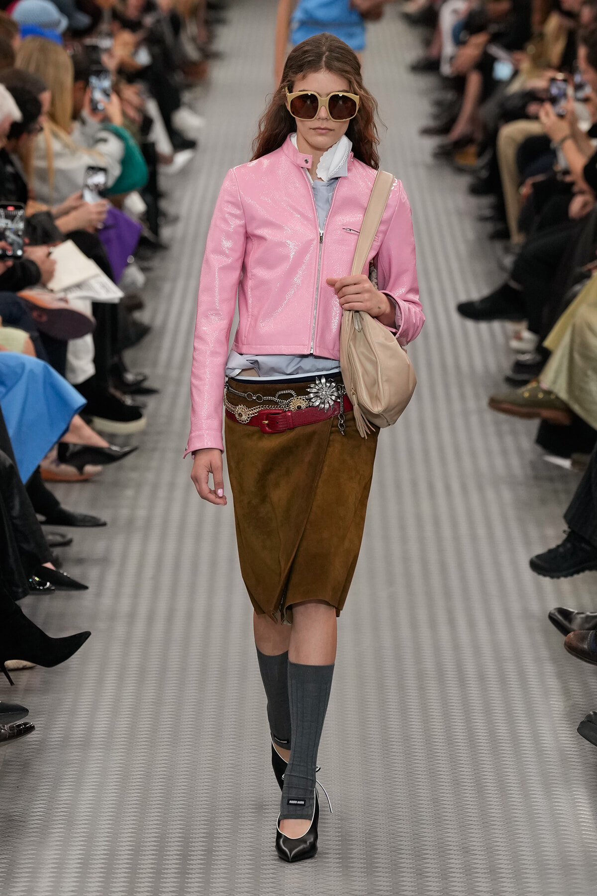 Model walks the runway in a pink patent jacket, brown suede skirt, gray knee-high socks, black heels, and a beige shoulder bag.