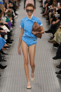 Model walking the runway in a light blue matching shirt and shorts, oversized sunglasses, and a brown handbag tucked under her arm.