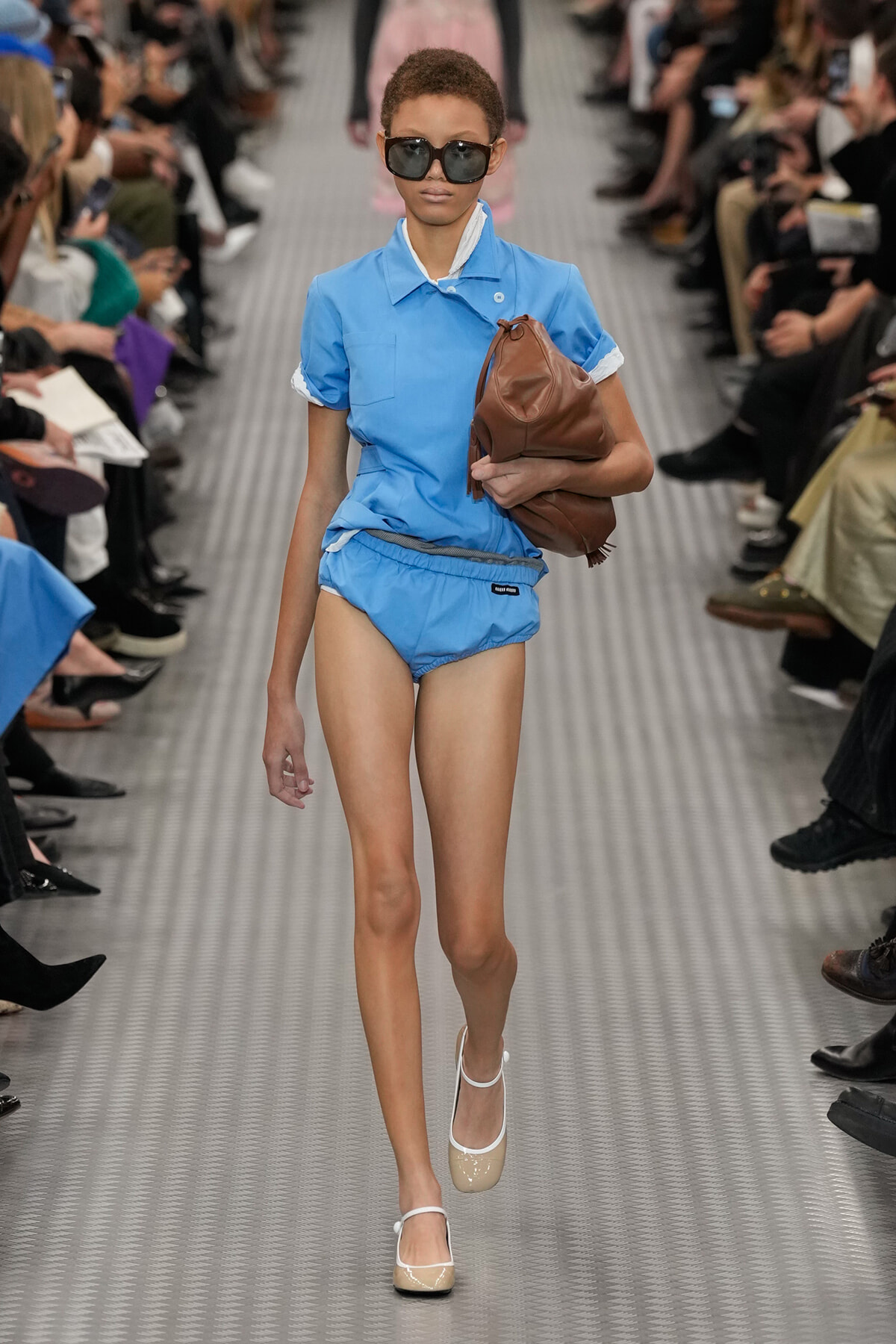 Model walking the runway in a light blue matching shirt and shorts, oversized sunglasses, and a brown handbag tucked under her arm.