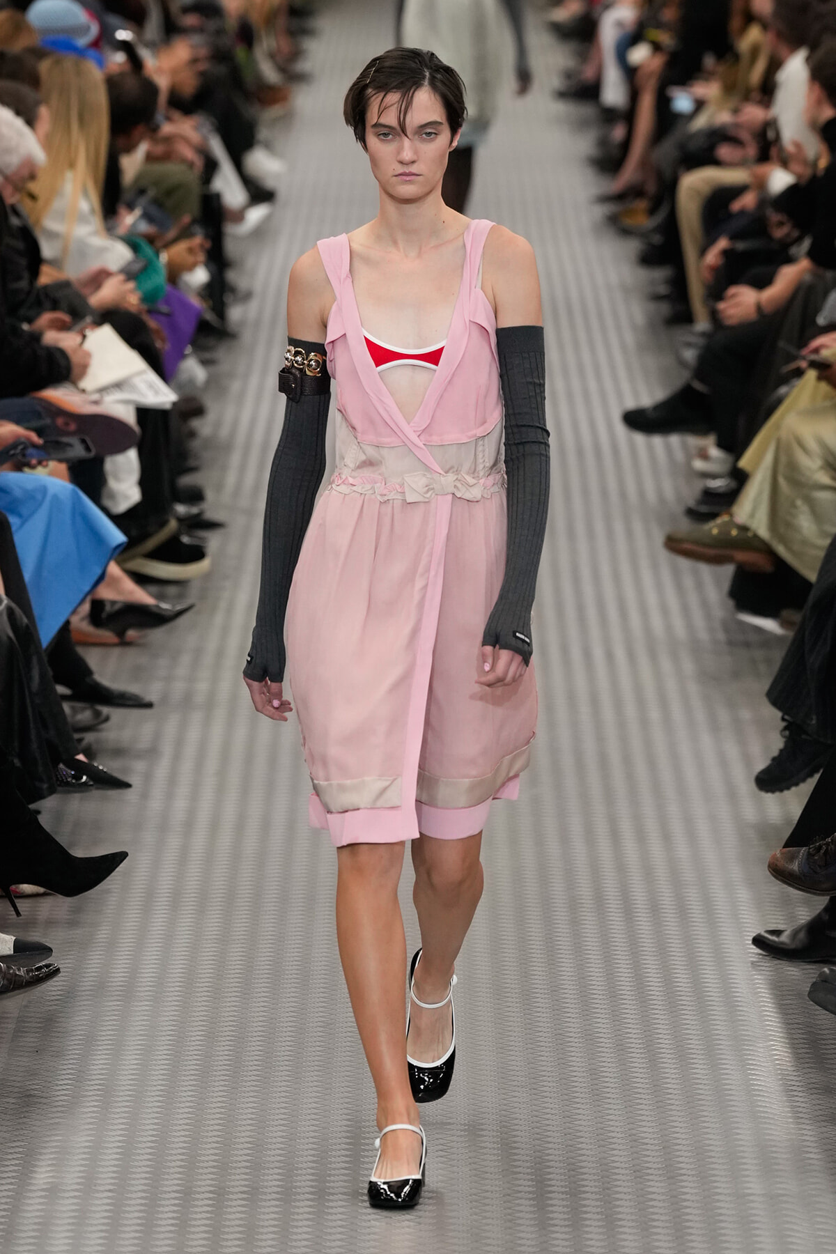 Model walks a runway in a pale pink layered dress with sheer waist, paired with gray arm warmers and black-and-white Mary Jane shoes.