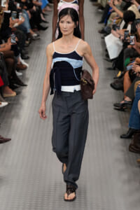 Model walking a fashion runway in a navy and light-blue strapped top, dark gray wide-leg pants, and brown handbag, on a metallic catwalk.