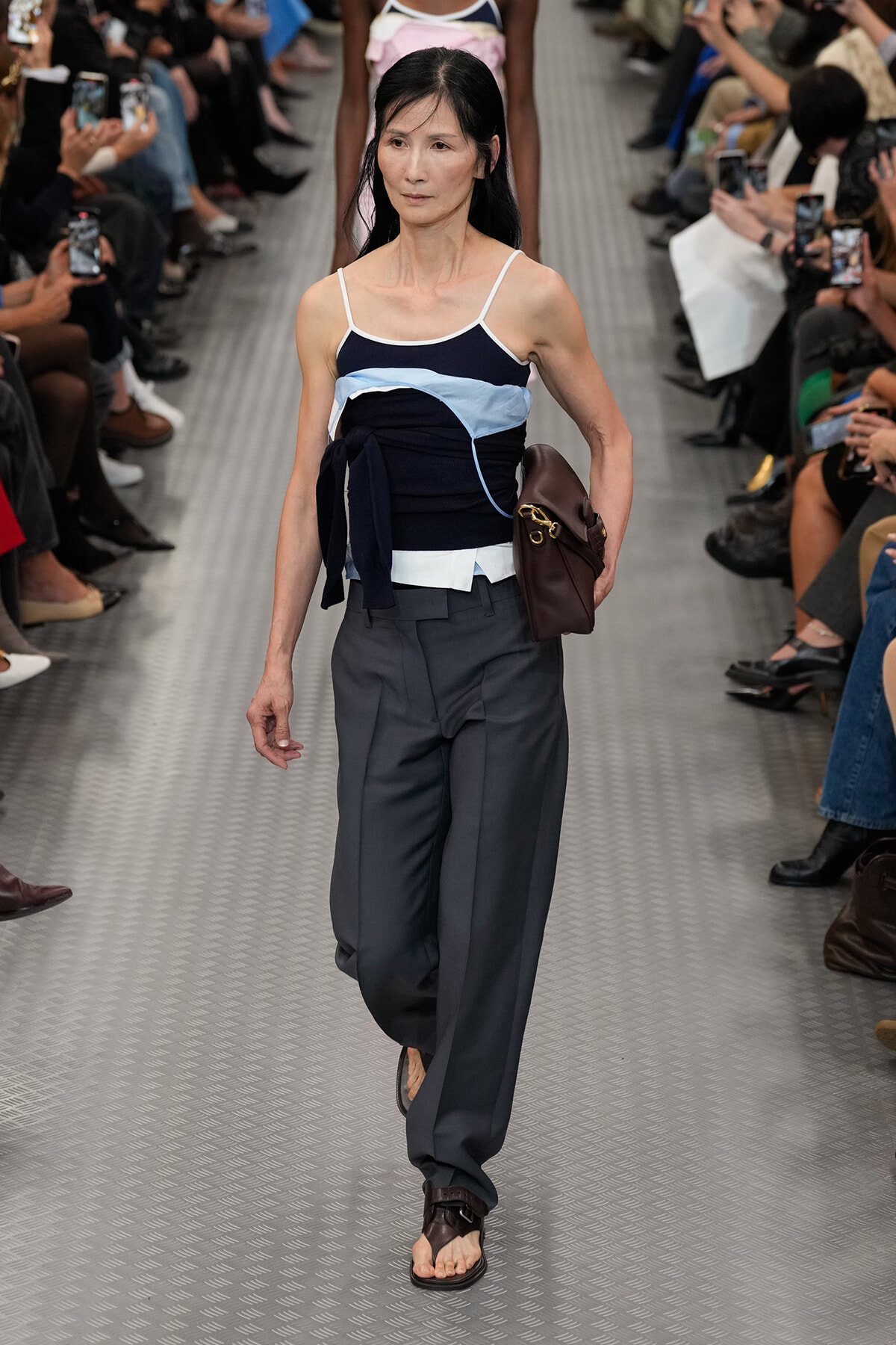 Model walking a fashion runway in a navy and light-blue strapped top, dark gray wide-leg pants, and brown handbag, on a metallic catwalk.