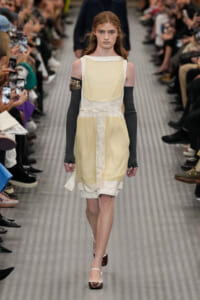 Model walking a fashion runway in a pale yellow layered dress with white trim and long black arm warmers, audience on sides.