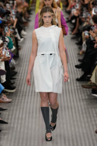 Female model on a fashion runway wearing a white sleeveless dress, knee-length, with gray knee-high socks and black open-toe heels.