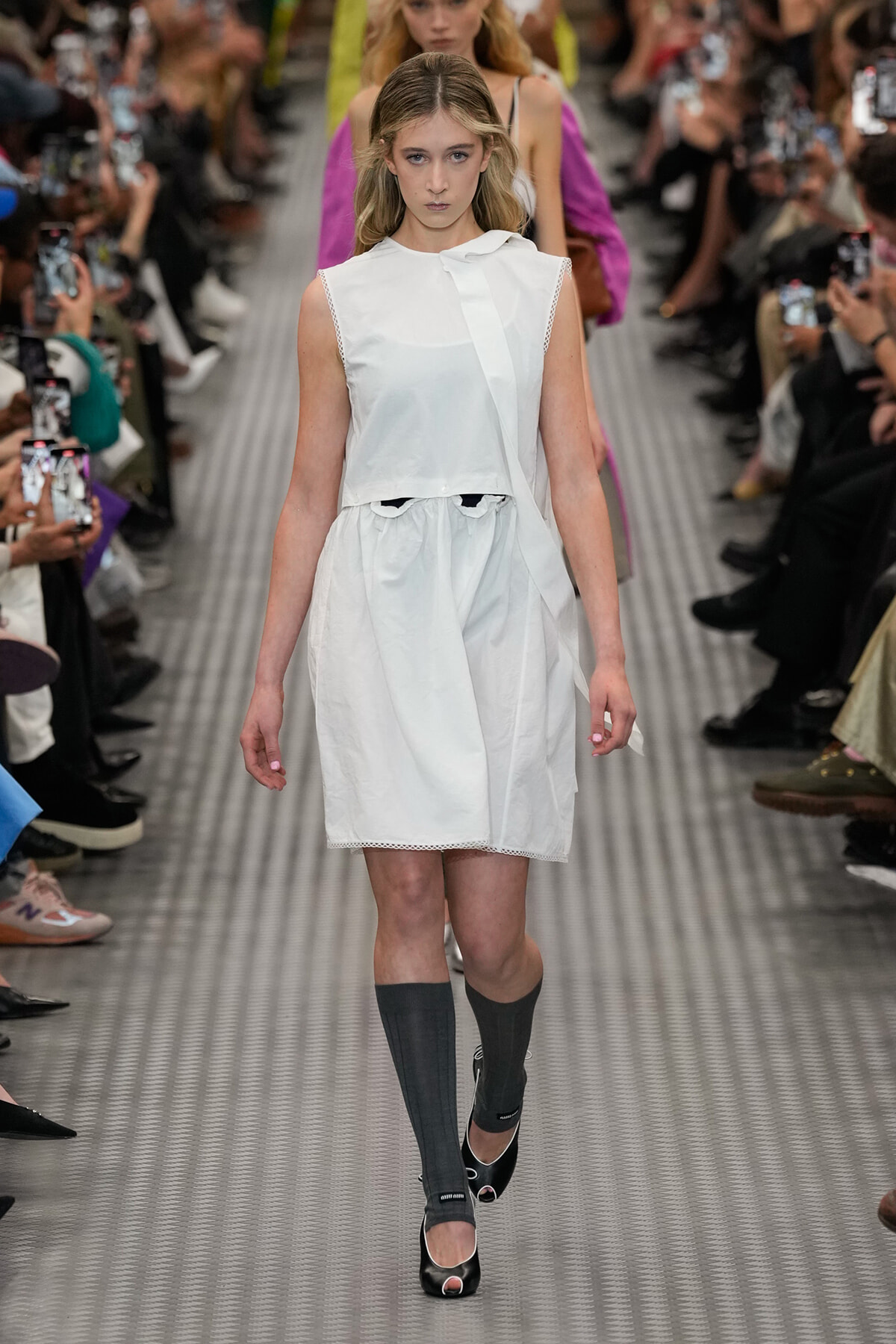 Female model on a fashion runway wearing a white sleeveless dress, knee-length, with gray knee-high socks and black open-toe heels.